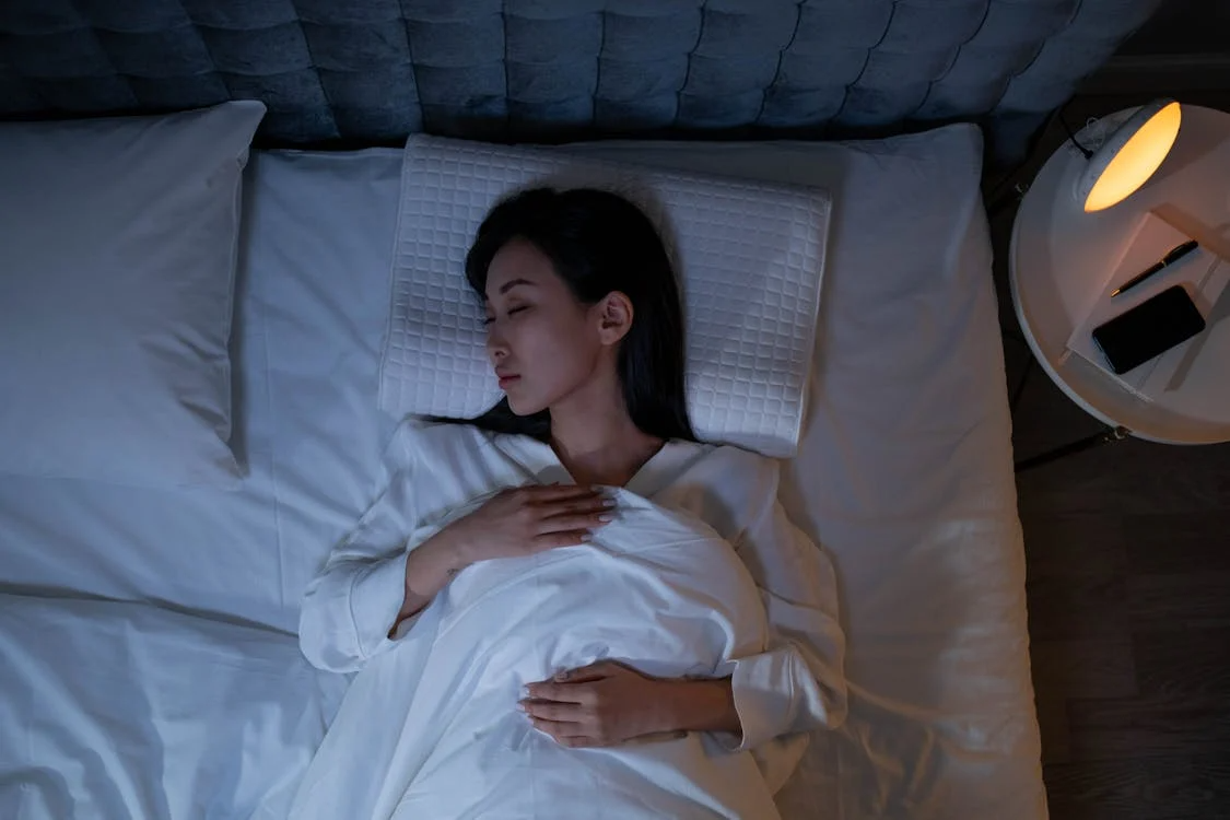 Woman sleeping on white bedding with warm bedside lamp, illustrating REM sleep and nighttime routine.